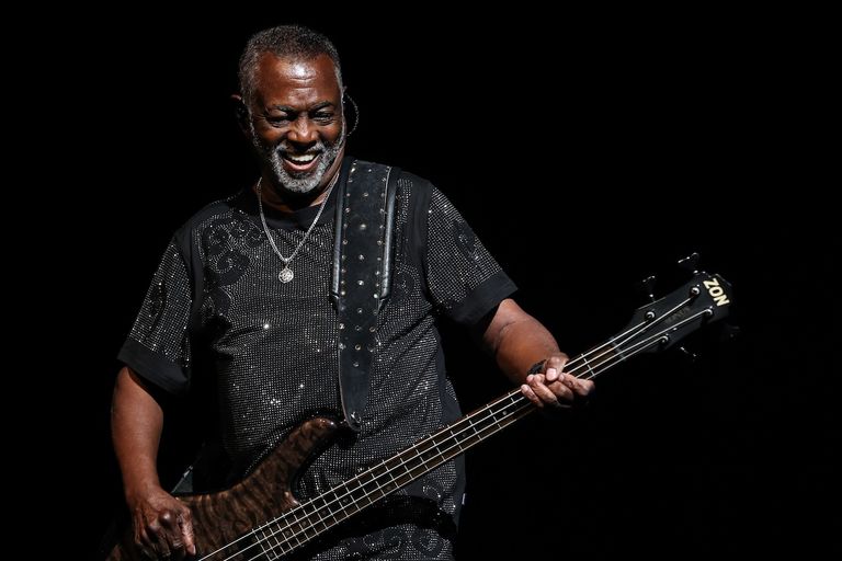 Robert "Kool" Bell, co-founder and bassist of Kool & The Gang, smiling in a black cap and t-shirt in front of a colorful backdrop.